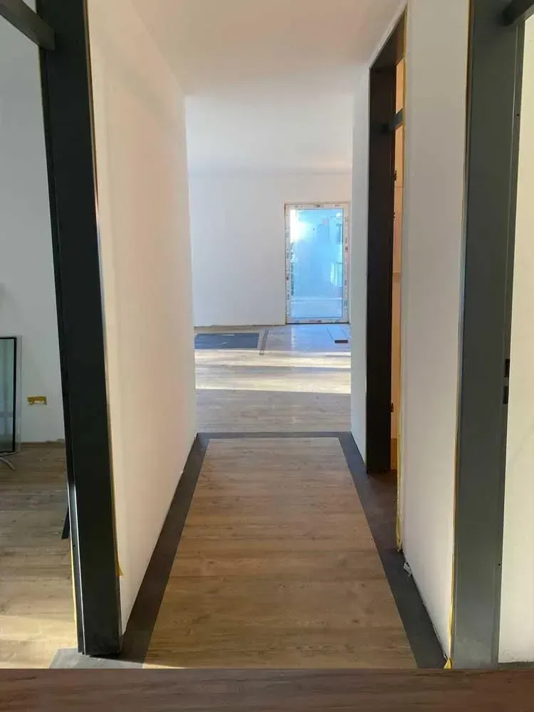 Schmaler Flur mit dunklen Türrahmen, Holzboden und einem hellen Zimmer am Ende