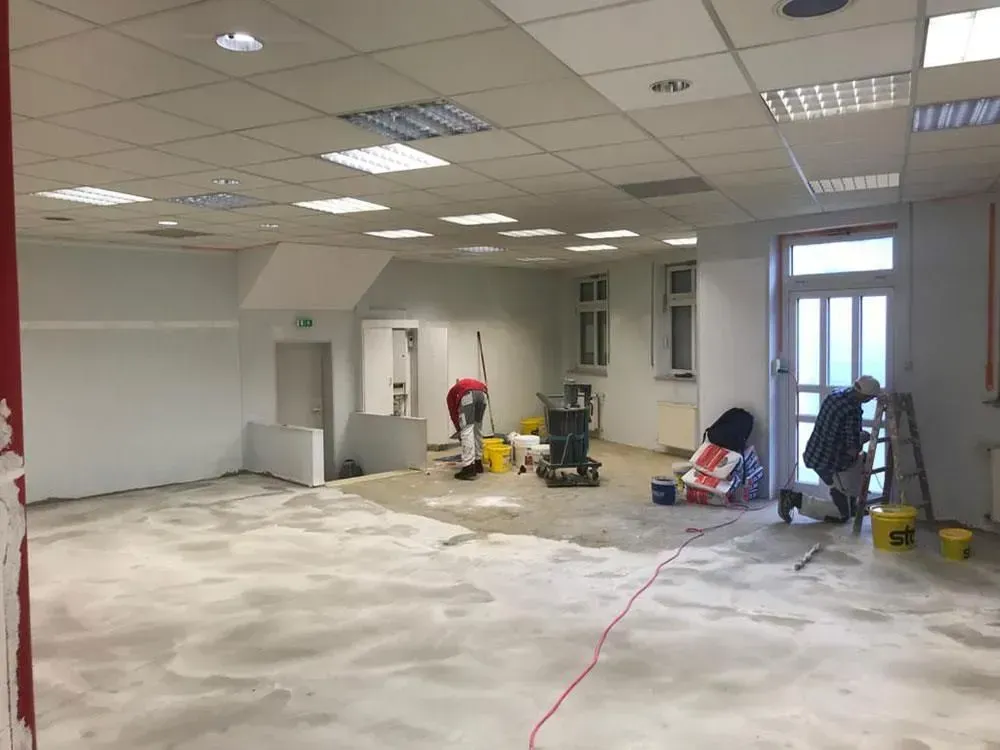 Ein leerer Raum, der gerade renoviert wird, mit Arbeitern, Werkzeugen und verstreuten Materialien auf dem Boden.