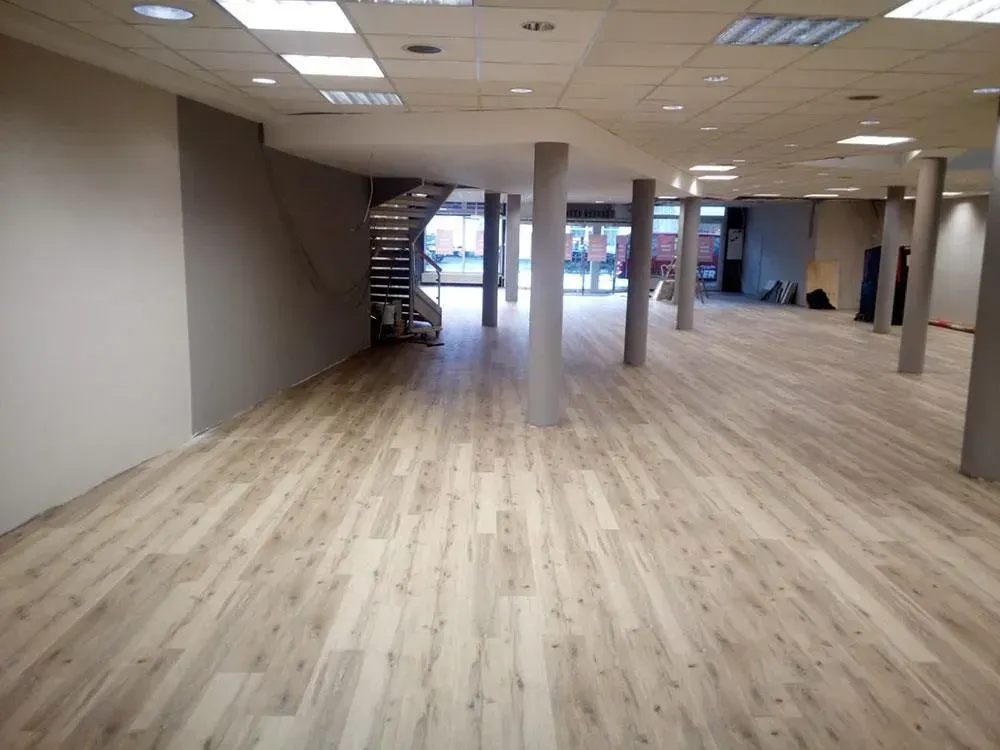 Leere, offene Bürofläche mit hellen Holzböden, weißen Deckenplatten und einer Treppe im hinteren Bereich.