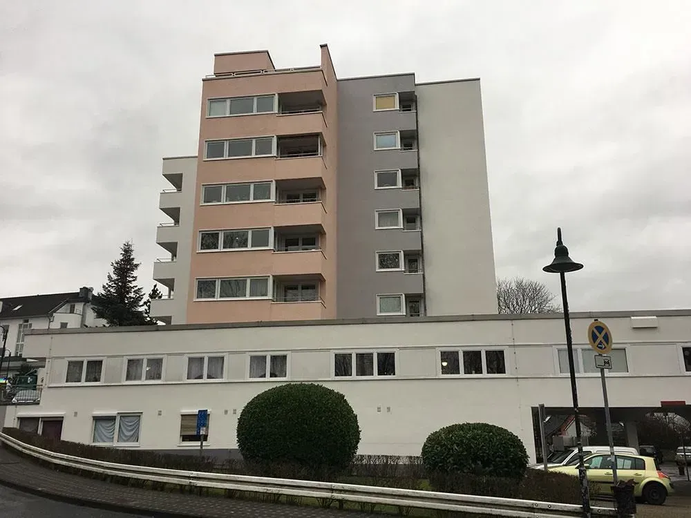 Wohnhaus unter bewölktem Himmel, davor ein weißes, niedrigeres Gebäudeteil, Büsche und geparkte Autos.
