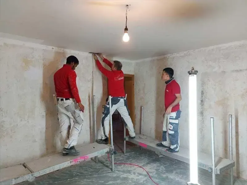 Drei Arbeiter in roten Hemden verputzen Wände in einem unfertigen Raum mit einer hängenden Glühbirne.