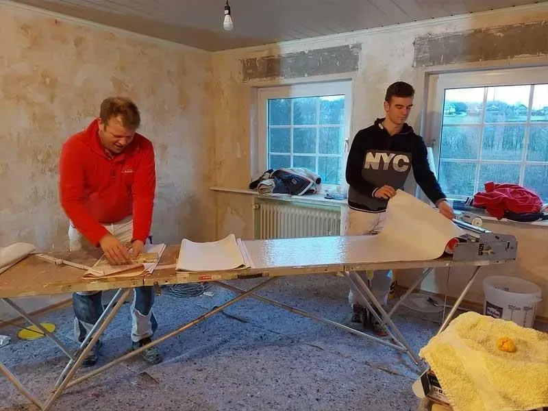 Zwei Personen tapezieren in einem spärlich möblierten Zimmer mit einer Leiter und Werkzeug.