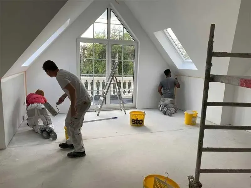 Arbeiter streichen einen Dachbodenraum mit weißen Wänden; Eimer und Leitern stehen in der Nähe eines großen Fensters.