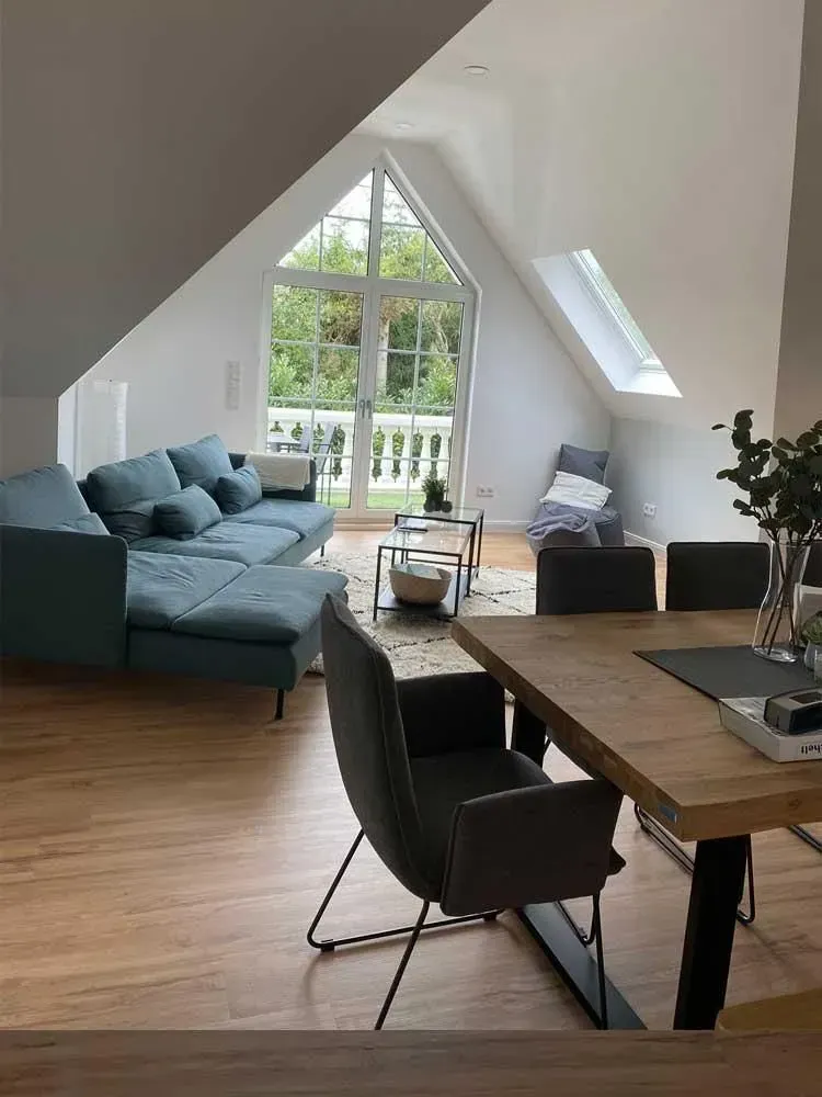 Helles Dachgeschosswohnzimmer mit blauem Sofa, Holztisch und großem dreieckigem Fenster mit Blick ins Grüne