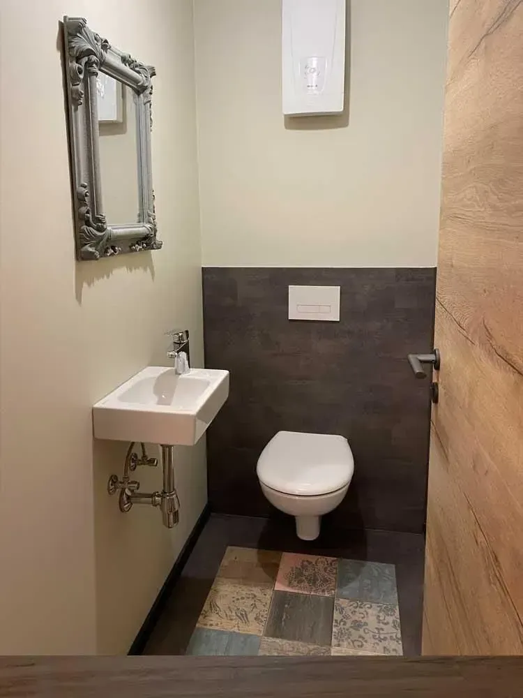 Kleine Toilette mit wandmontiertem WC, winzigem Waschbecken, Spiegel und mehrfarbigem Teppich.