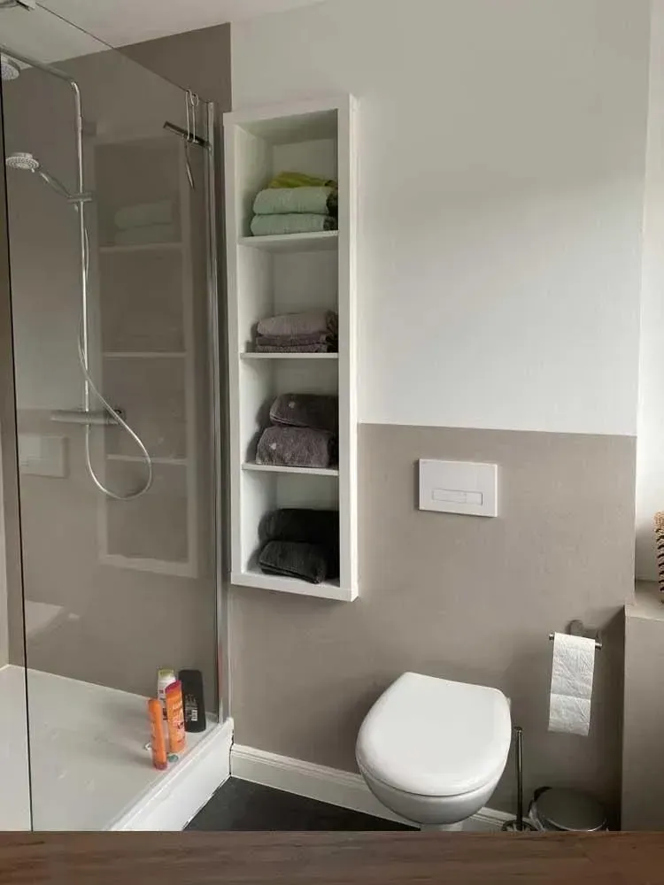 Modernes Badezimmer mit Dusche, Toilette und eingebautem Regal zur Aufbewahrung gefalteter Handtücher.