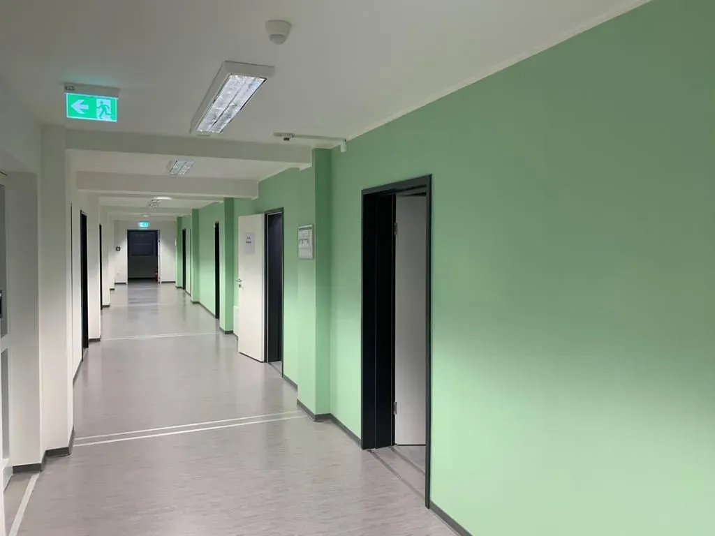 Leerer mintgrüner Flur mit offenen Türen und Neonröhren an der Decke.