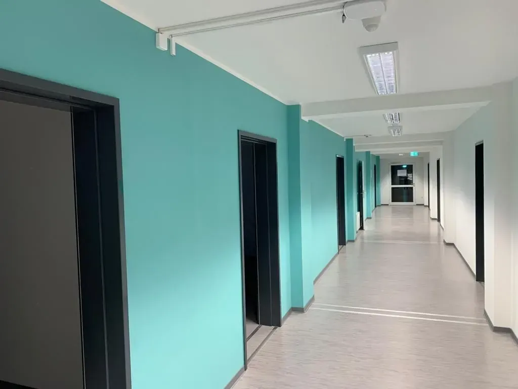 Leerer Krankenhausflur mit türkisfarbenen Wänden, weißem Boden und offenen Türen unter Neonlicht