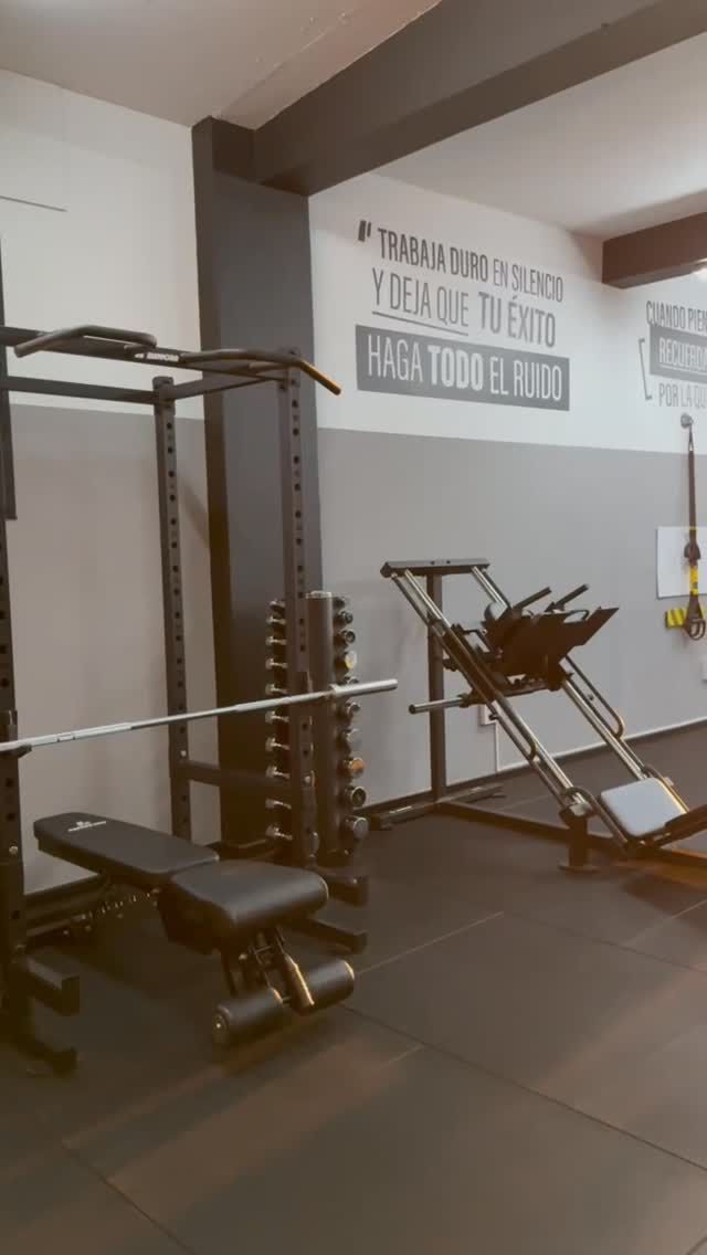 Gimnasio vacío con banco de pesas, soporte para pesas y equipo de ejercicio contra paredes blancas.