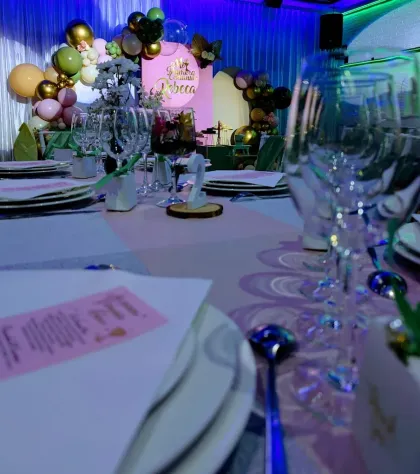 Elegante mesa de banquete con platos y cristalería blancos, frente a un escenario decorado con globos bajo iluminación azul.