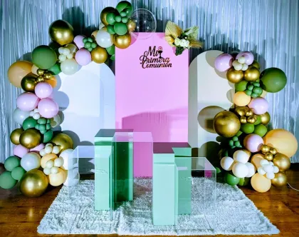 Arco de globos en tonos pastel y dorados enmarcando un fondo rosa con mesas de cristal color menta sobre una alfombra blanca.