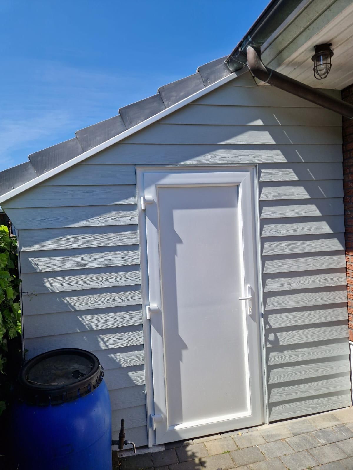 An der Seite eines Gebäudes neben einem blauen Fass befindet sich eine weiße Tür.