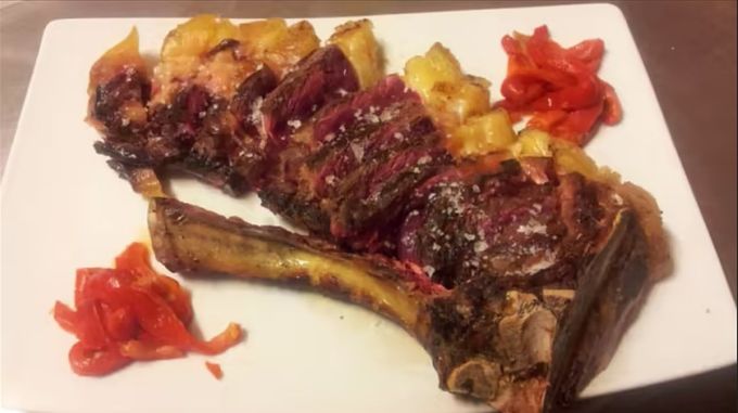 Bistec a la parrilla con hueso, patatas y pimientos rojos en un plato blanco.