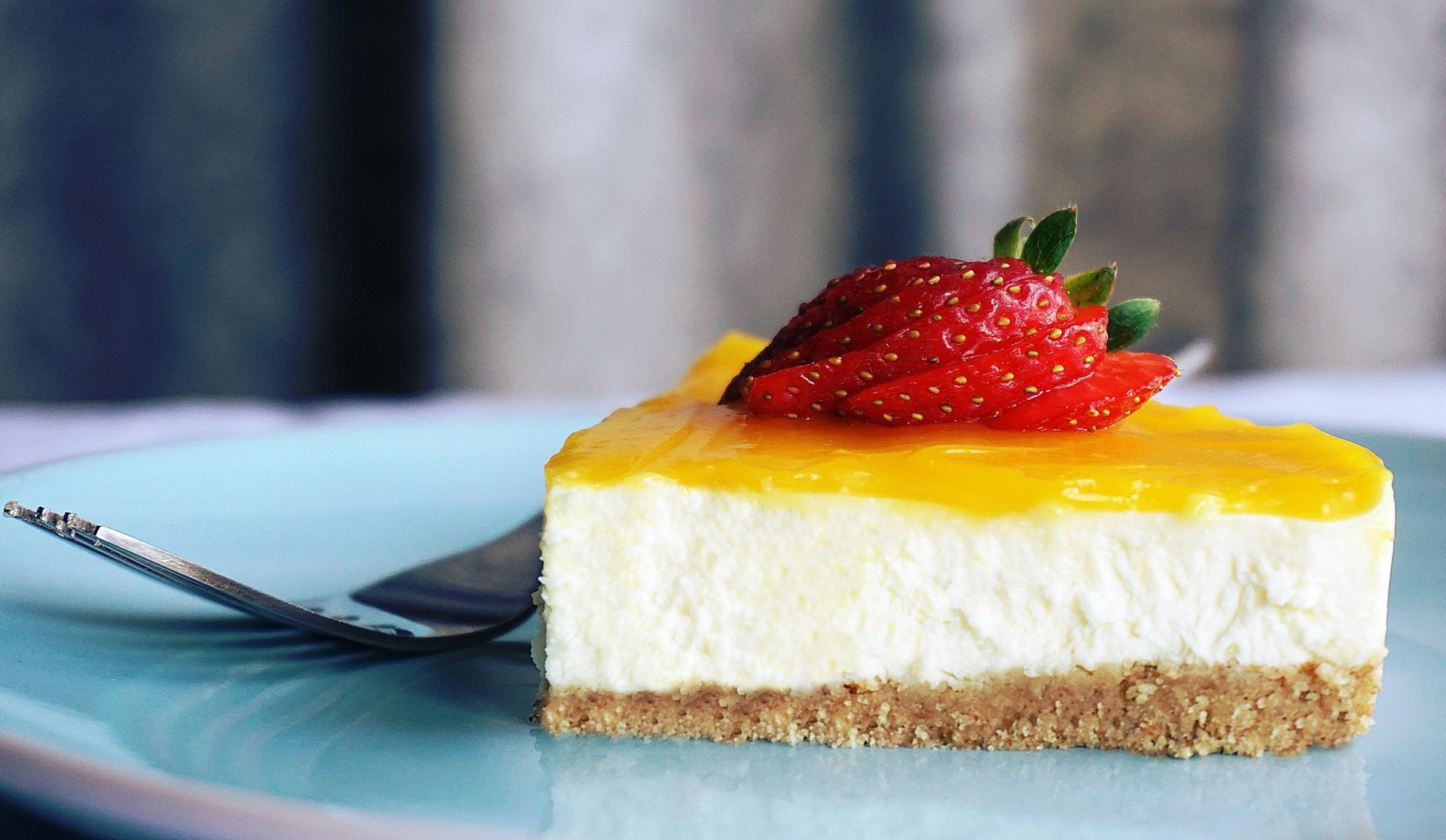 Cuadrado de tarta de queso cubierto con glaseado amarillo y fresa en un plato azul con un tenedor.