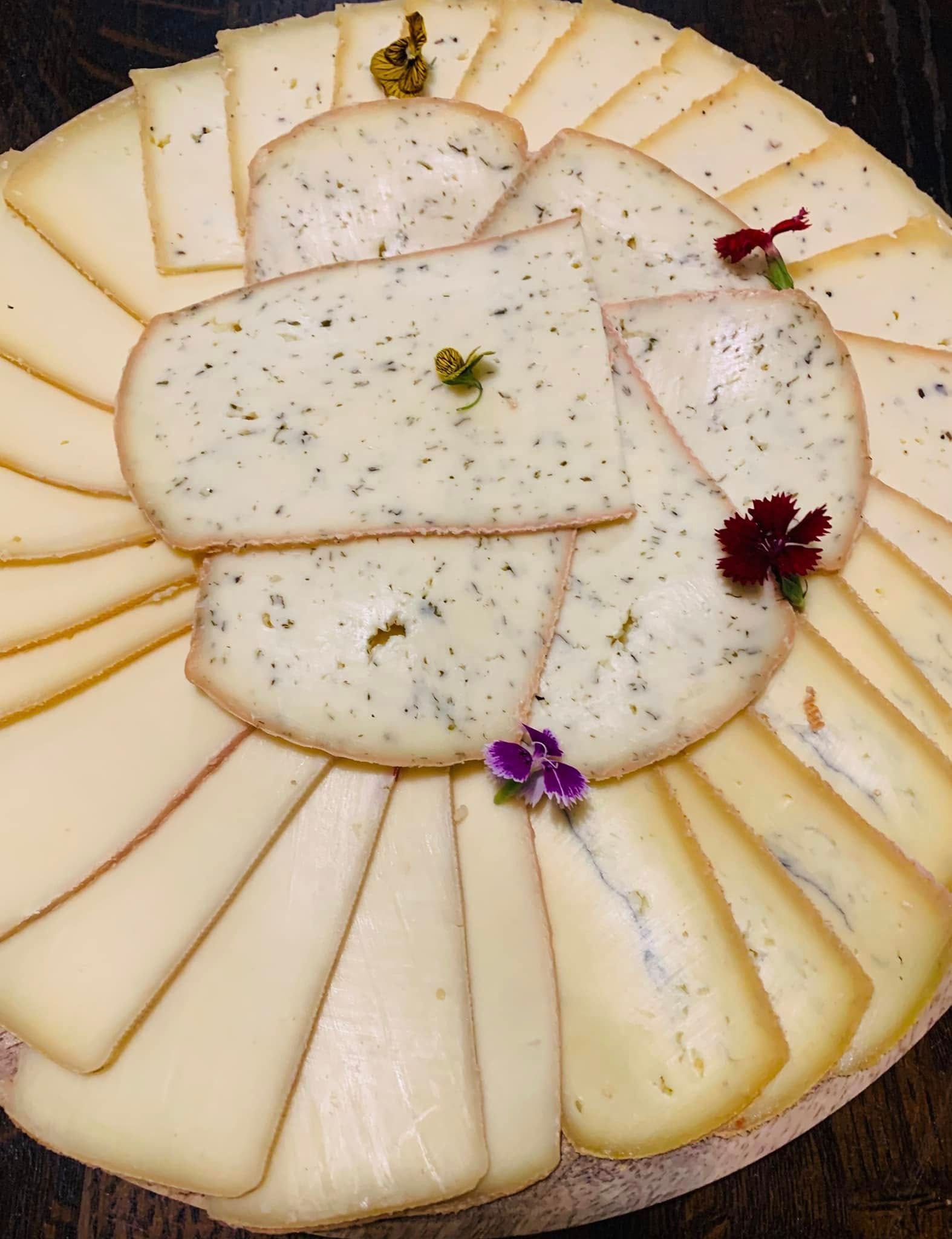 Plateau de fromages finement tranchés et d'herbes aromatiques, garni de petites fleurs.