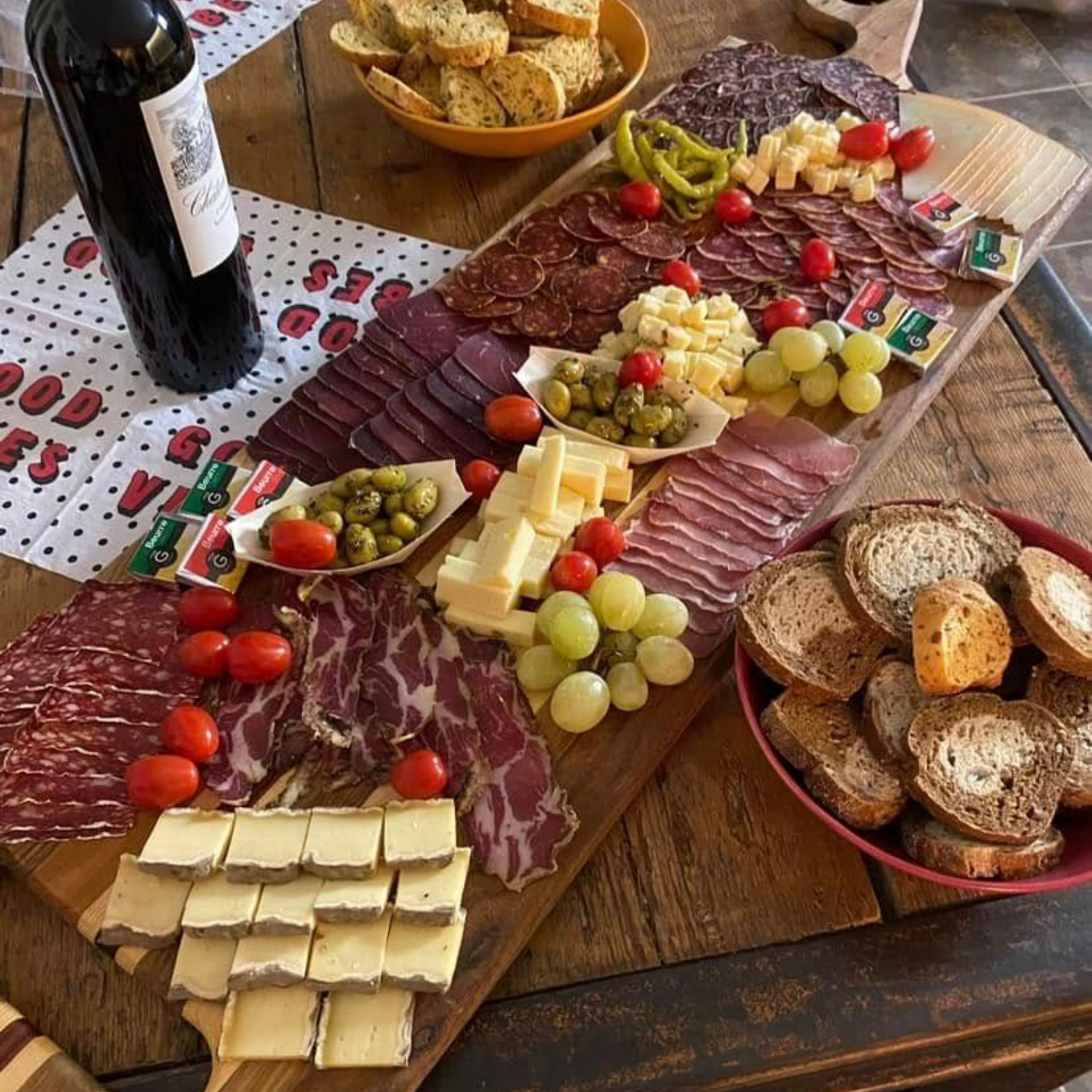 Planche de charcuterie avec viandes, fromages, olives, raisins et crackers, à côté d'une bouteille de vin sur une table.