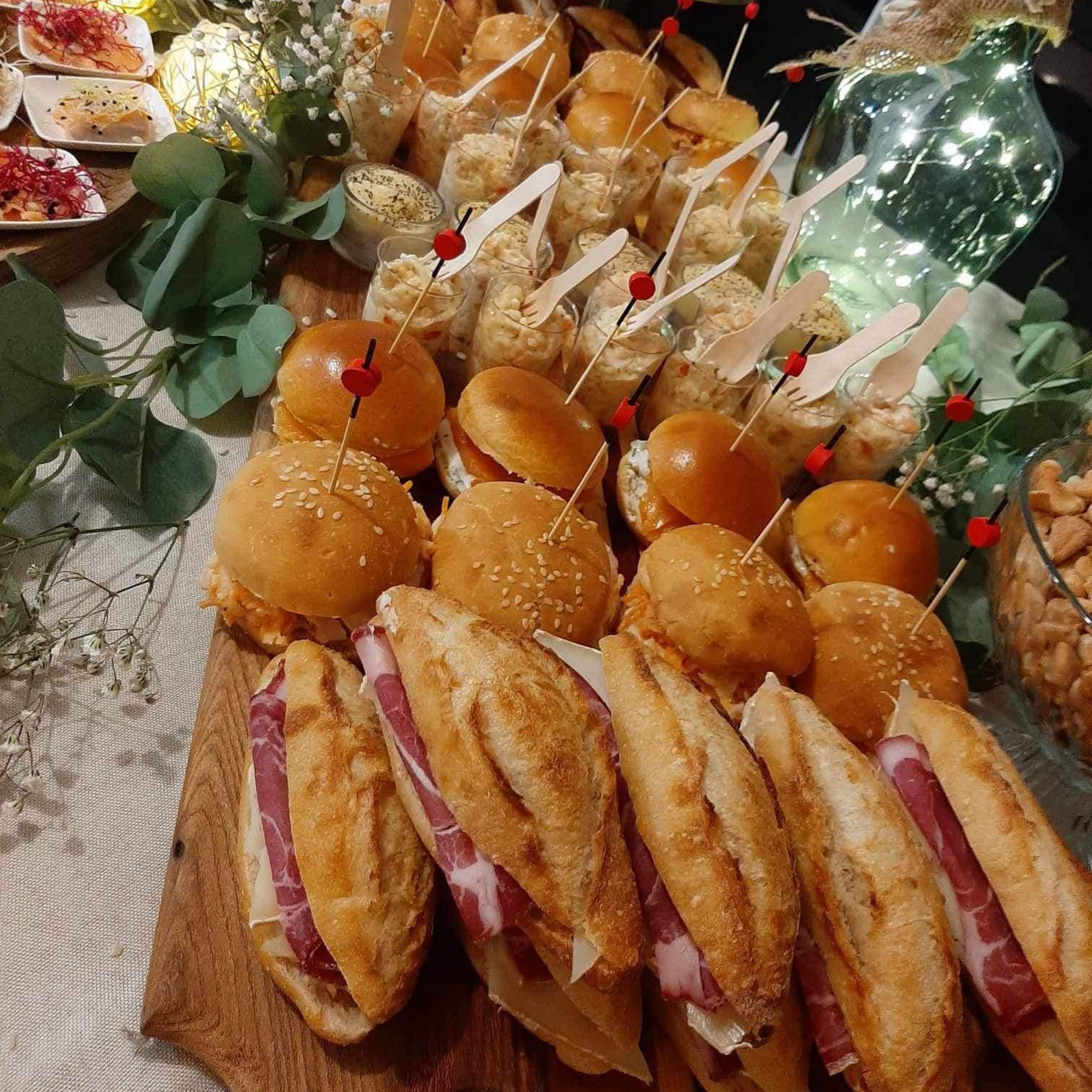 Plateau traiteur composé de sandwichs assortis, de miniburgers et d'amuse-gueules présentés sur une planche en bois.