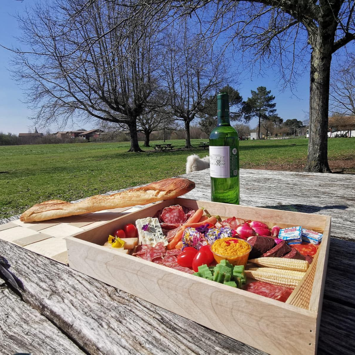 Pique-nique sur une table en bois à l'extérieur : baguette, planche de charcuterie et bouteille de vin dans un parc ensoleillé.