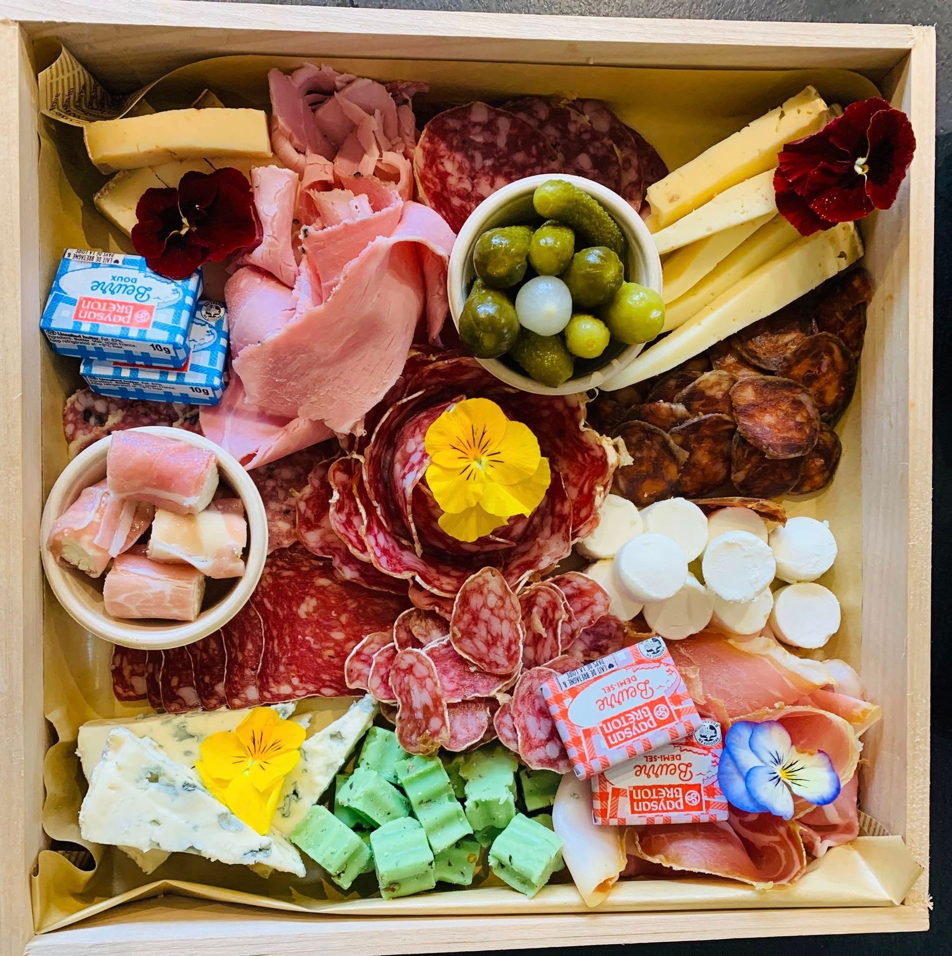Planche de charcuterie : fromages, charcuteries, olives, cornichons, fleurs, dans une boîte en bois.