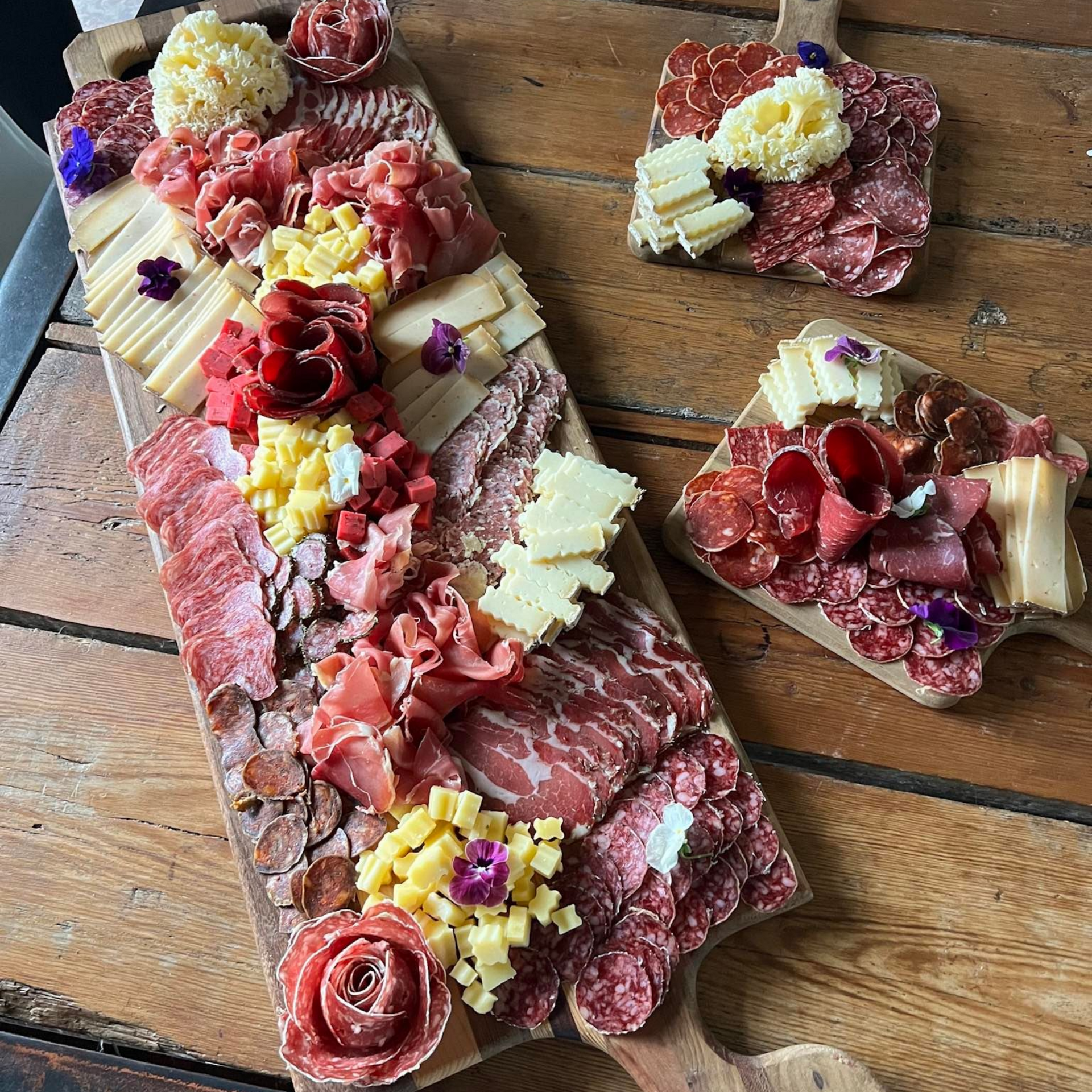 Planches de charcuterie sur une table en bois : fromages, viandes et fleurs disposés de façon décorative.
