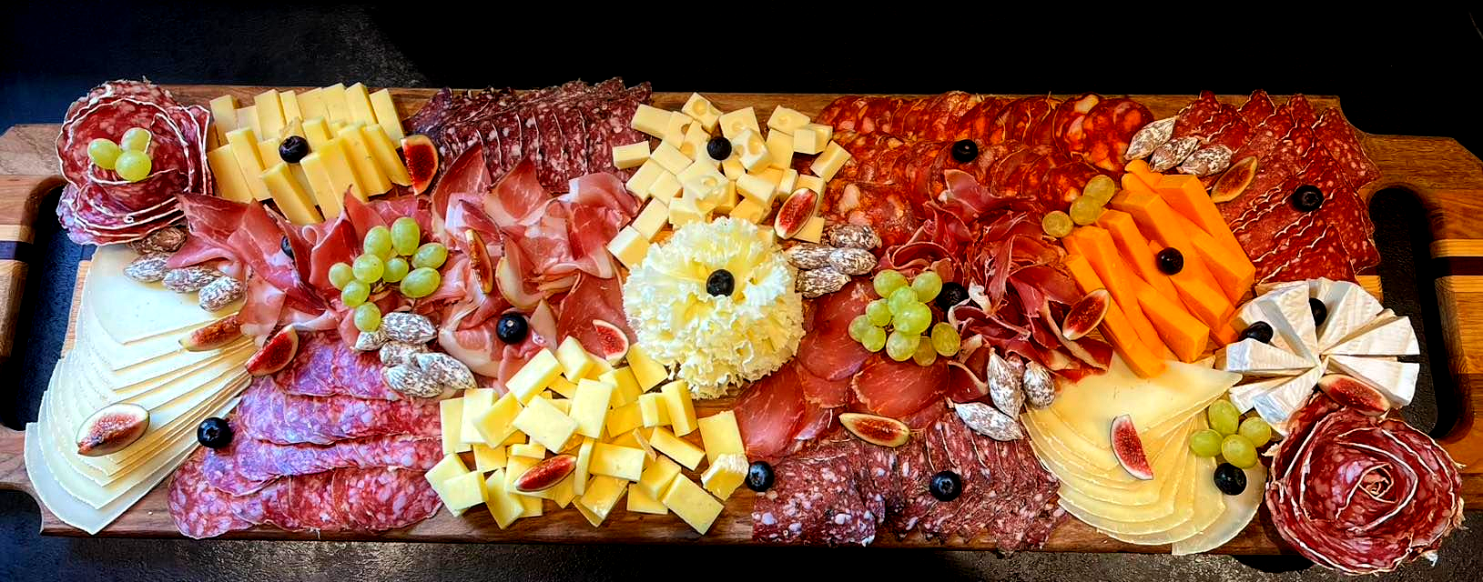Planche de charcuterie composée de viandes variées, de fromages, de raisins et de garnitures décoratives, présentée sur un plateau de service en bois.