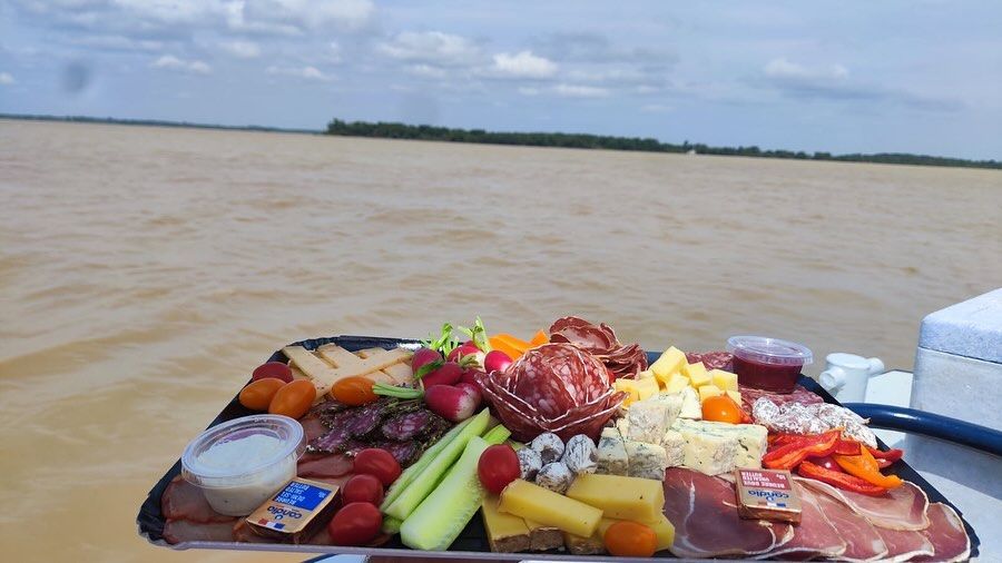 Planche de charcuterie avec viandes, fromages et fruits sur un bateau, avec vue sur une rivière.