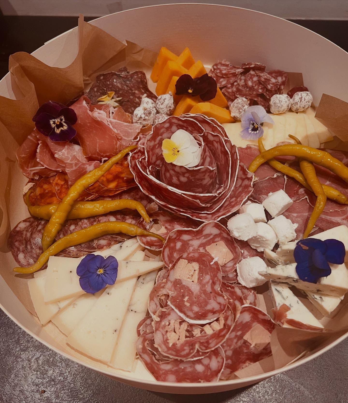 Planche de charcuterie composée de viandes, de fromages, de poivrons et de fleurs disposés dans un récipient rond.