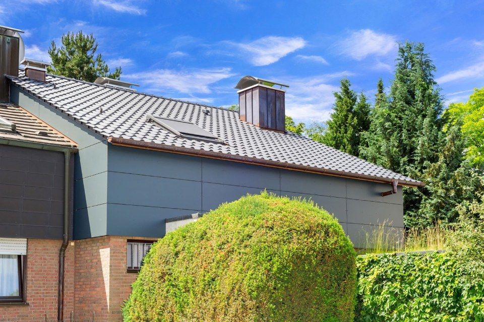 HB Holzbau Breymaier GmbH – graue Hausfassade mit hellgrauem Schrägdach hinter einer Hecke