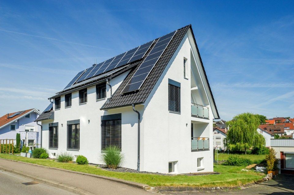 HB Holzbau Breymaier GmbH – kontrastreiches Foto eines weißen Mehrfamilienhauses mit schwarzen Dachschindeln und Solarpaneelen vor blauem Himmel