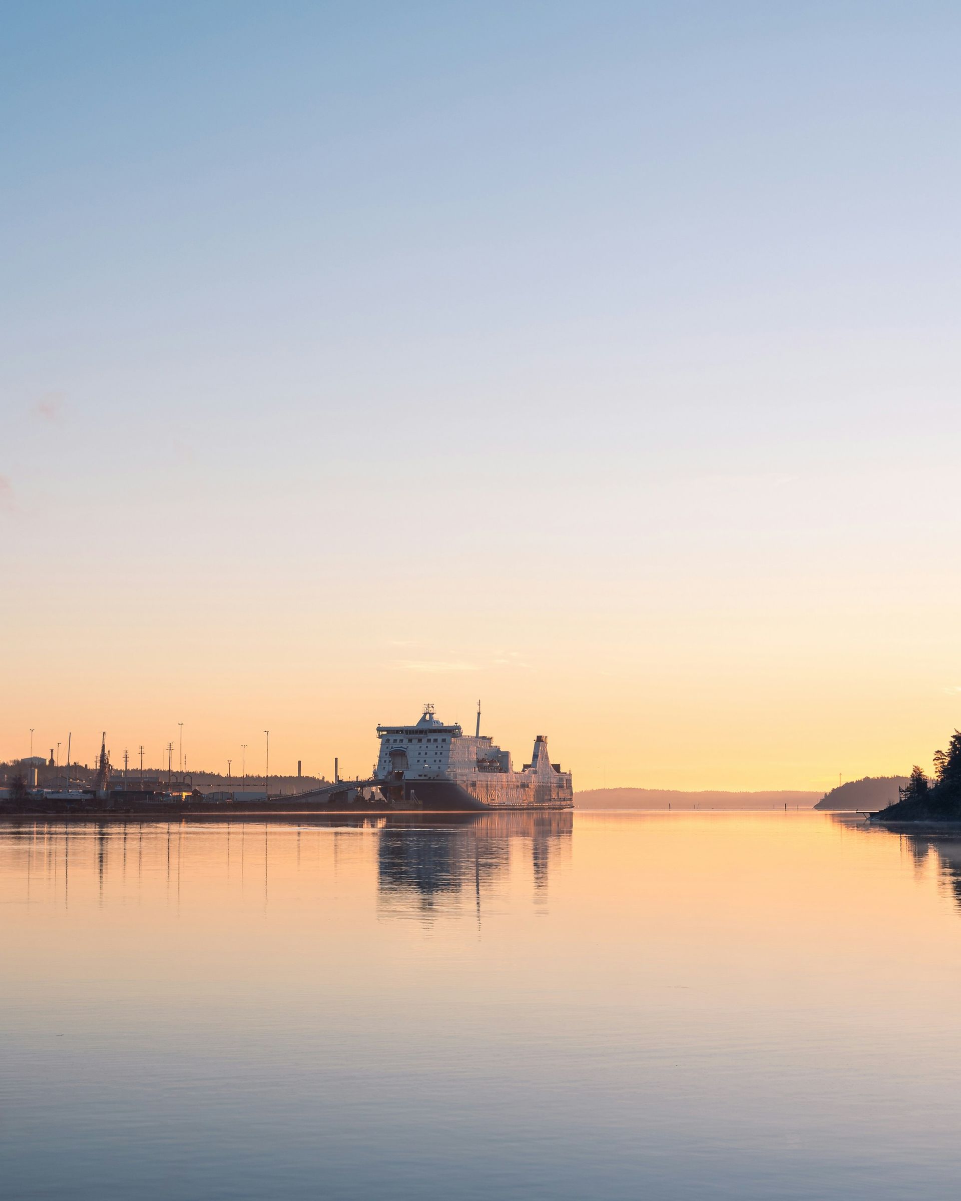 Calm harbor at sunrise with a docked ship silhouetted against a pastel sky and mirror-like water.
