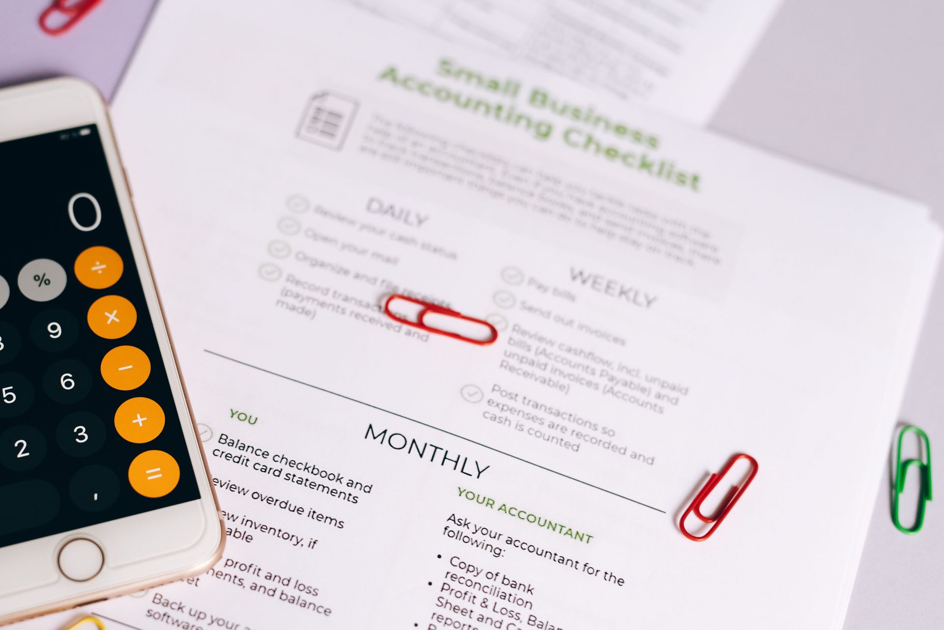 Smartphone calculator beside a small business accounting checklist with red and green paper clips