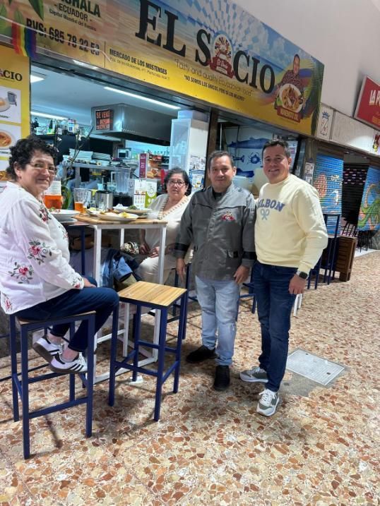 Cuatro personas, de pie y sentadas, en un puesto de comida del restaurante El Socio, con un cartel amarillo