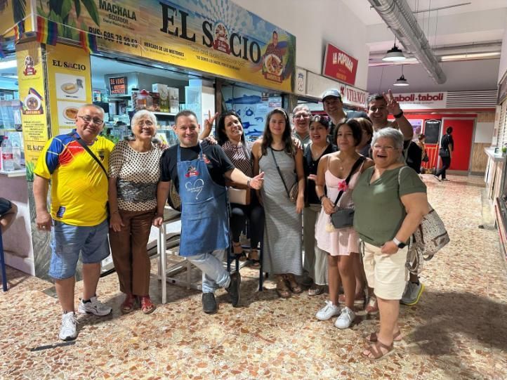 Un grupo diverso de personas sonríe para una foto frente a un puesto de mercado llamado El Socio; todos visten ropa informal.