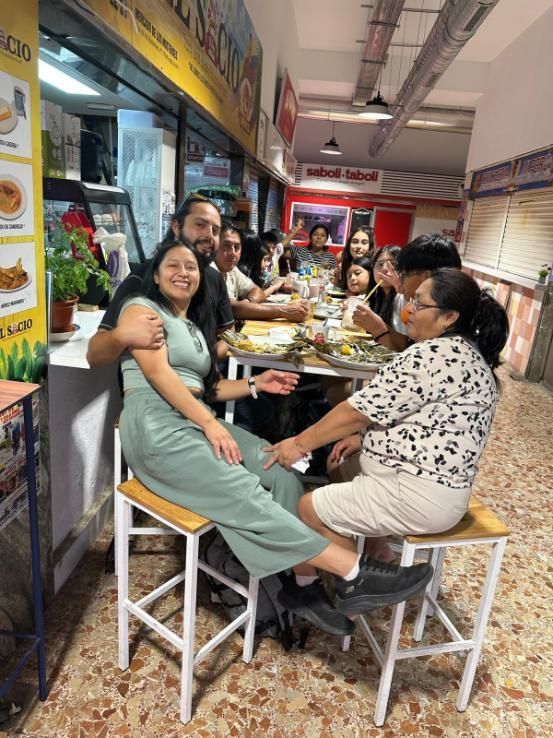 Un grupo de personas se sienta en mesas en un patio de comidas bien iluminado, sonriendo y charlando mientras comen juntas.