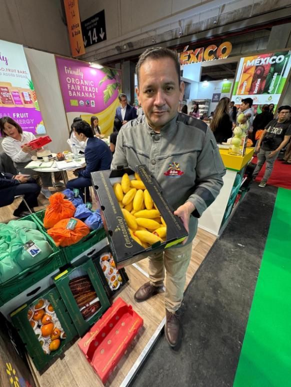 Una persona se encuentra en un stand de una feria comercial, sosteniendo una caja de mangos amarillos