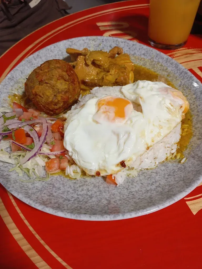 Un plato que incluye dos huevos fritos sobre arroz, una albóndiga empanizada, pollo al curry y salsa fresca.