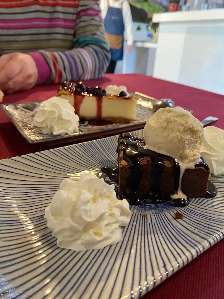 Postres de tarta de queso y brownie en platos estampados, con crema batida y helado, sobre un mantel rojo.