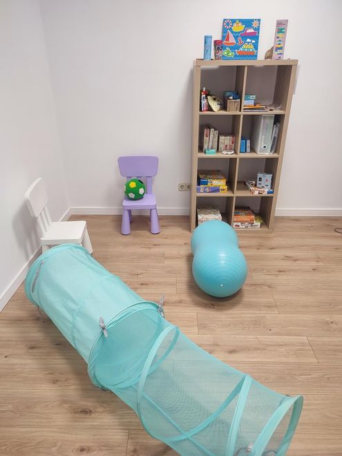 Un área de juegos con un túnel azul, dos sillas, una pelota y una estantería con juguetes.