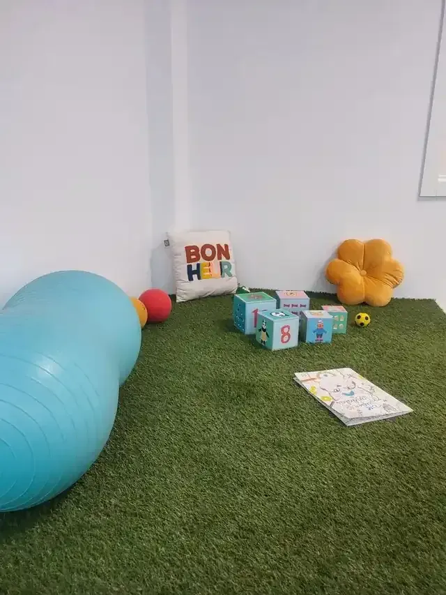 Sala de juegos con alfombra verde, pelota de ejercicio azul, bloques de juguete, almohada y cojín en forma de flor.