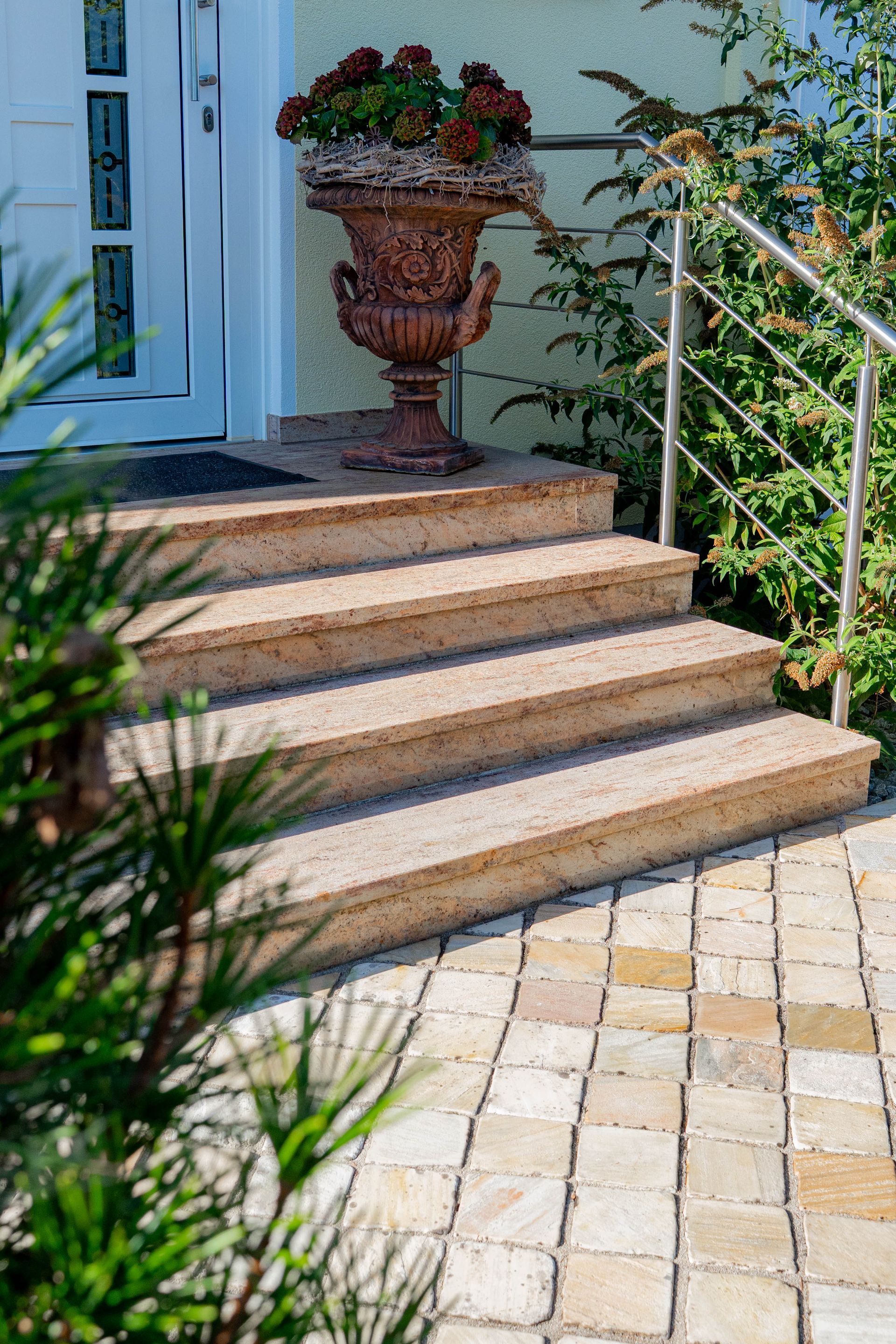 Natursteintreppen mit einem Stahlhandlauf zu einer Haustür und vielen Pflanzen und mediterranen Dekorationen im Vorgarten 