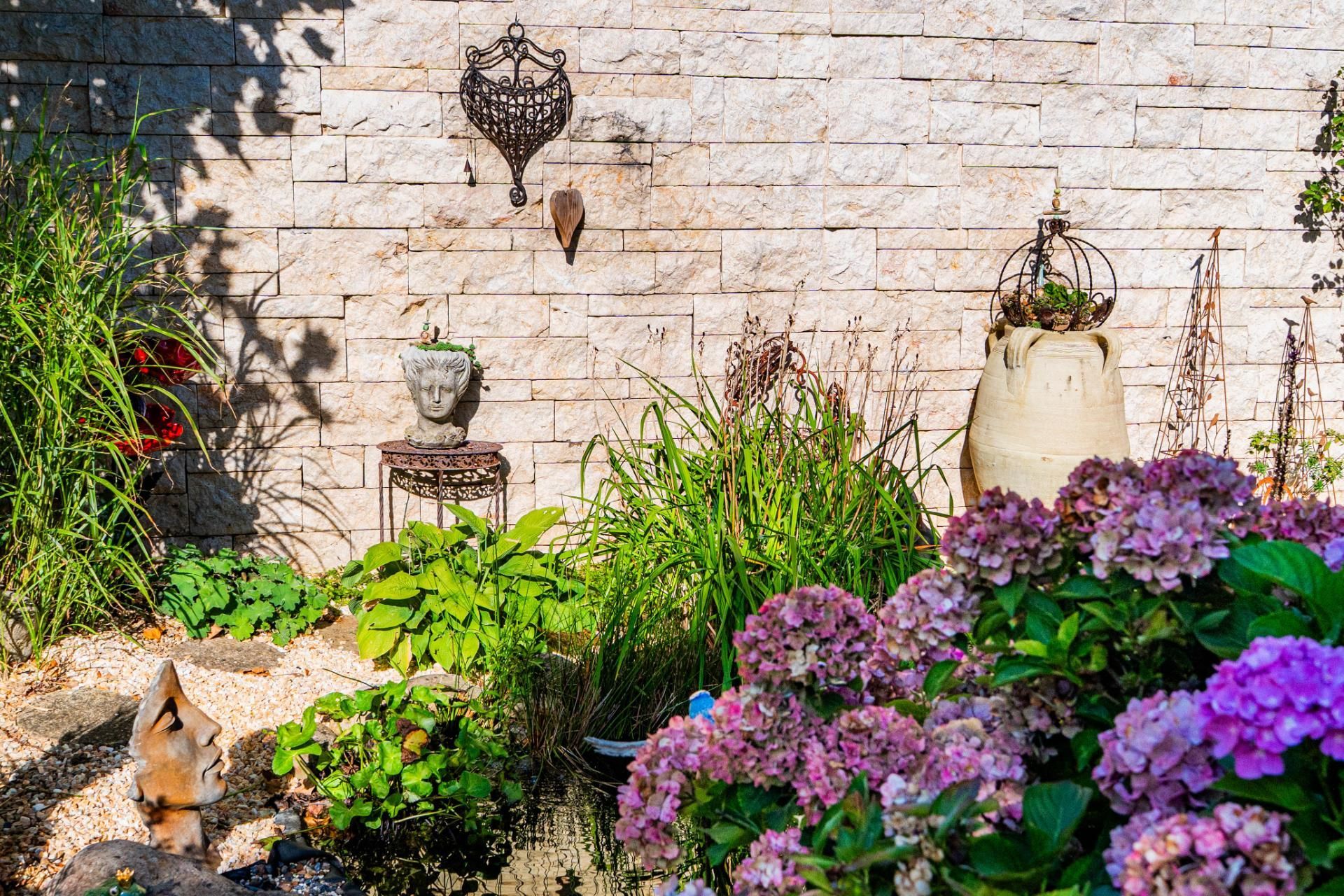 Garten mit vielen bunten Pflanzen, einer Natursteinwand als Sichtschutz und Skulpturen/Deko