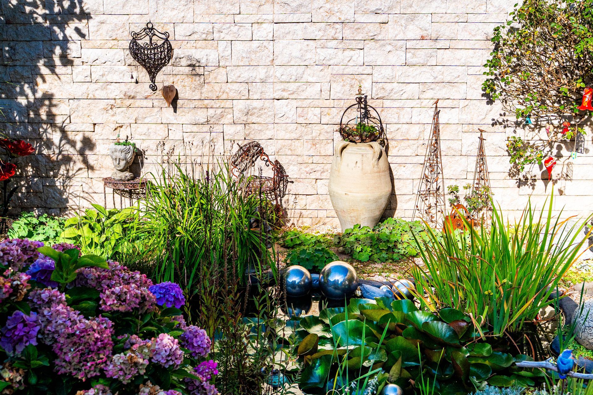 Garten mit vielen bunten Pflanzen, einer Natursteinwand als Sichtschutz, Skulpturen/Deko und einem kleinen Teich