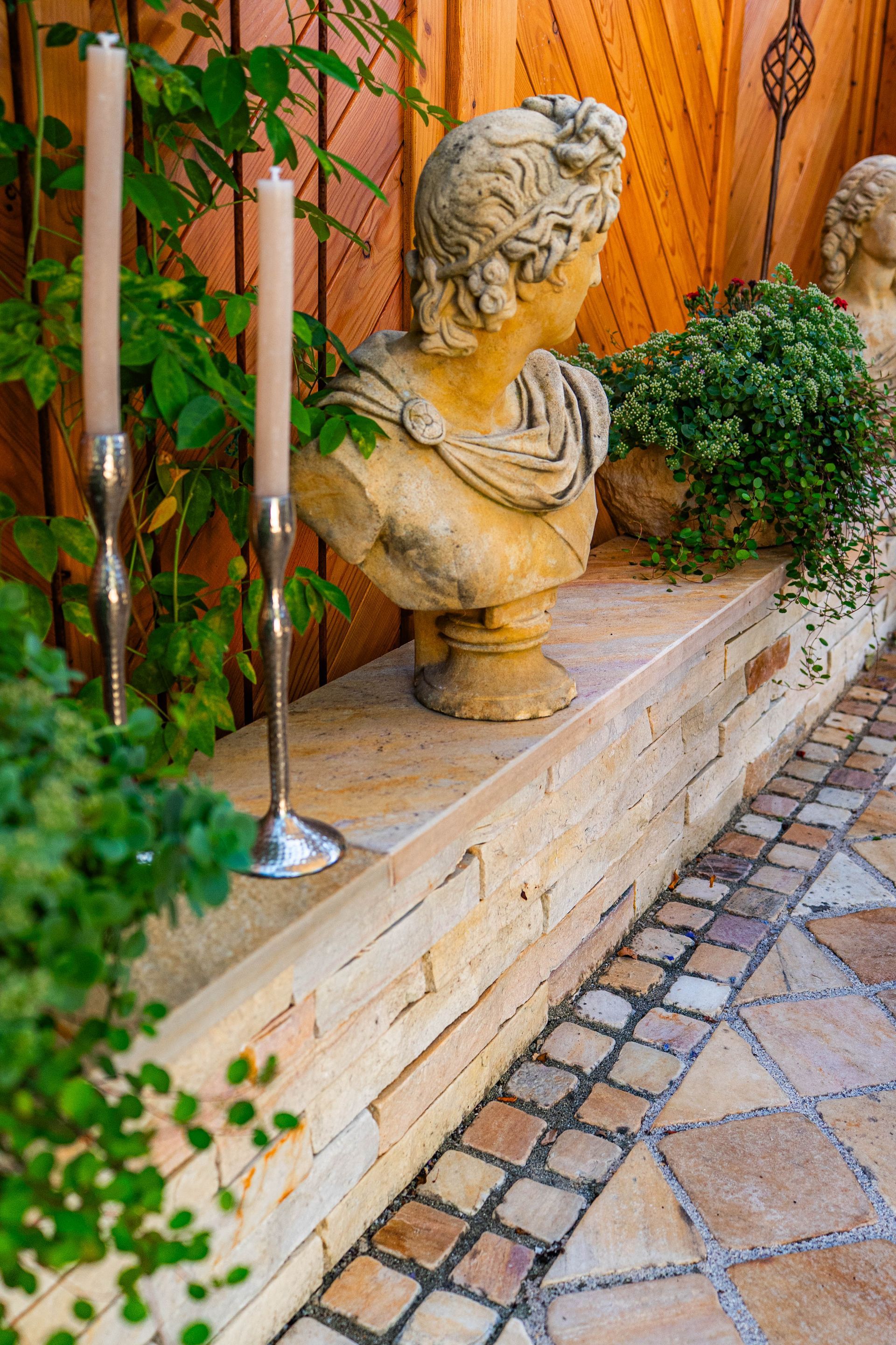 Garten mit einem Sichtschutz-Holzzaun und einer langen Steinbank mit Pflanzen, Stabkerzen und einer Skulptur