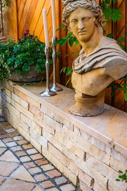 Garten mit einem Sichtschutz-Holzzaun und einer langen Steinbank mit Pflanzen, Stabkerzen und einer Skulptur