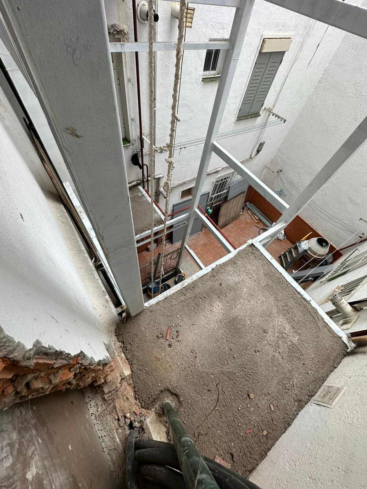 Mirando hacia abajo desde una estrecha escalera metálica gris en un pozo exterior. Se ven una pared de ladrillo, tuberías y pequeños pisos.