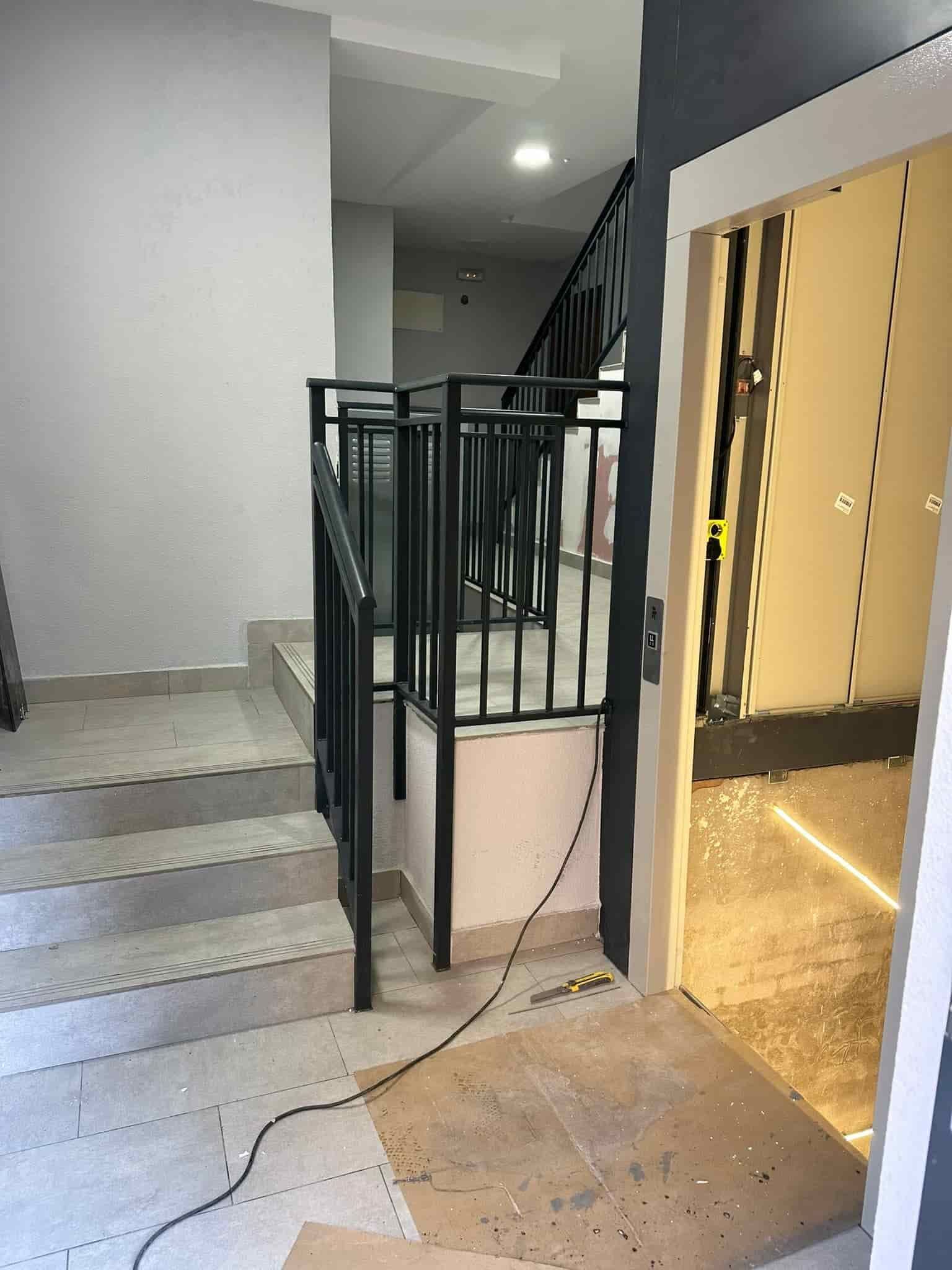 Escalera con barandilla metálica de color negro y entrada a ascensor en pasillo.