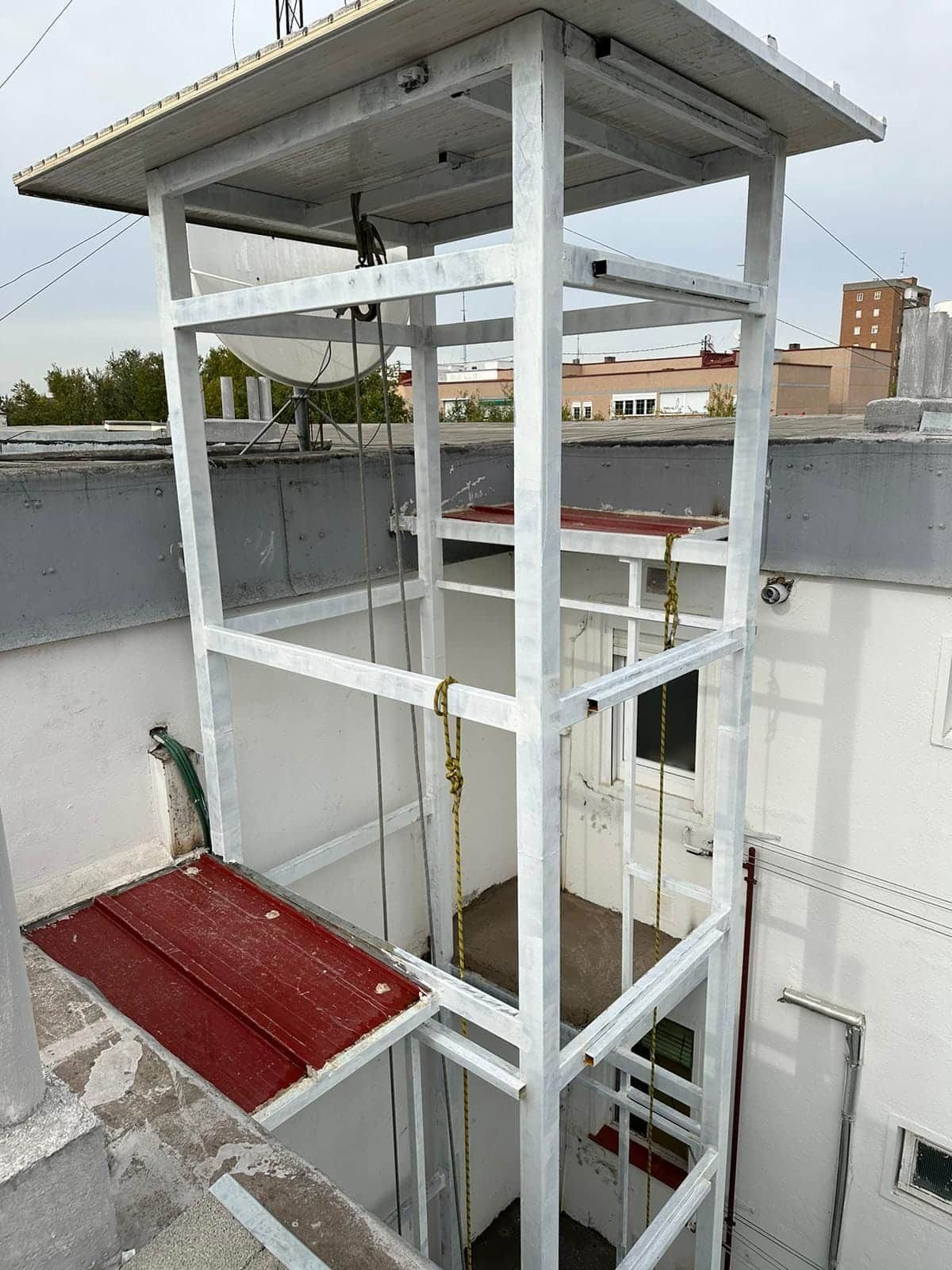 Estructura de ascensor de techo de metal blanco; plataforma roja; cables visibles.