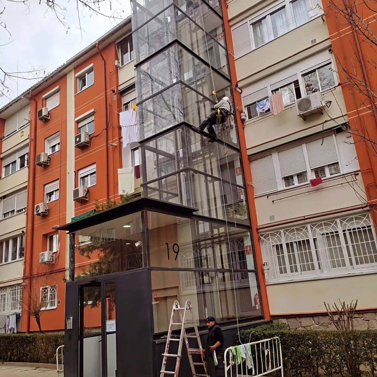 Ascensor exterior de cristal adosado a un edificio naranja y beige de varias plantas. Un trabajador está suspendido en su interior.