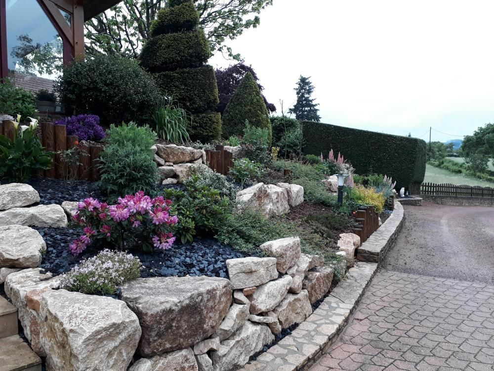 Jardin de rocaille avec des fleurs roses et des arbustes taillés, au bord d’une route pavée
