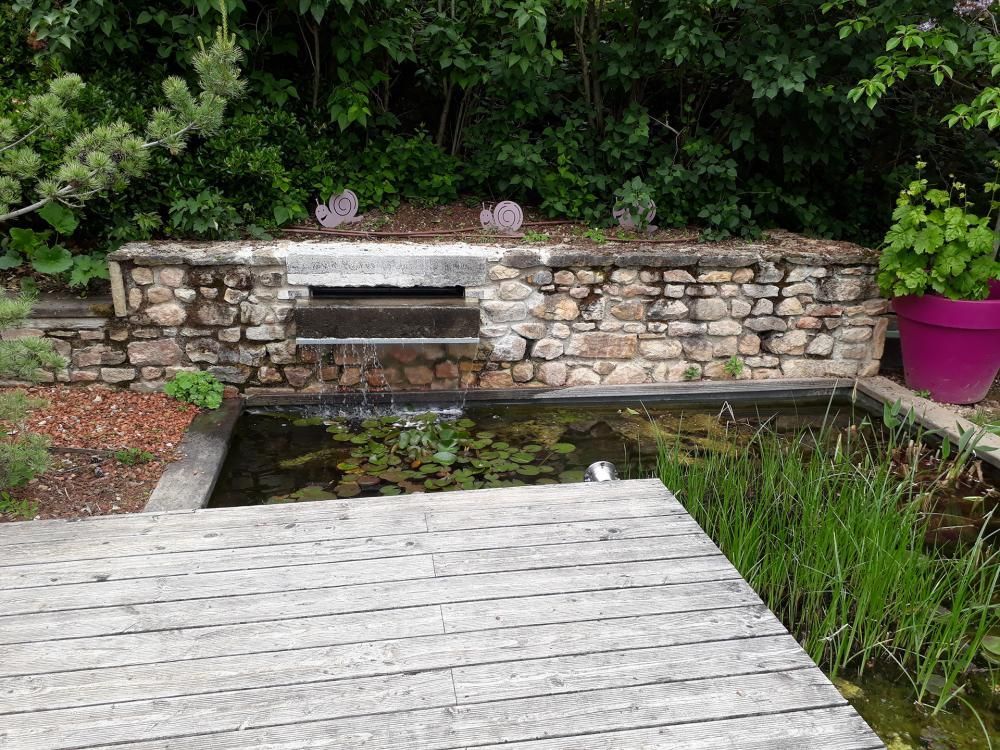 Mur en pierre au bord d’un petit bassin, avec une terrasse en bois blanc et de la verdure tout autour
